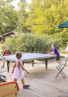 Niños y adultos juegan al ping-pong al aire libre en un parque vacacional en Île de France rodeados de árboles.