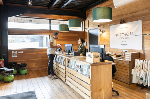 Recepción de Huttopia Versailles con un huésped y personal, decoración de madera y folletos turísticos.