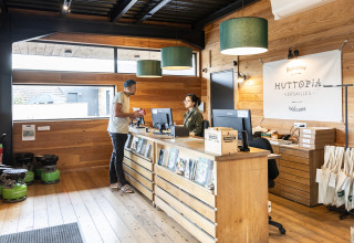 Receptie van Huttopia Versailles vakantiepark met klant en medewerker, houten aankleding en folders.