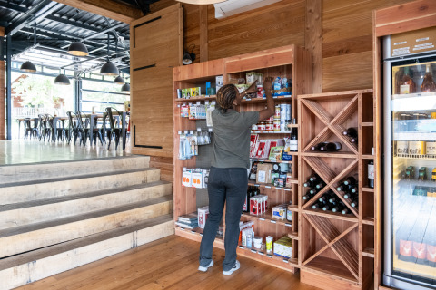 Persona organizando productos en una estantería de madera en una tienda, con botellas de vino y bebidas cerca.
