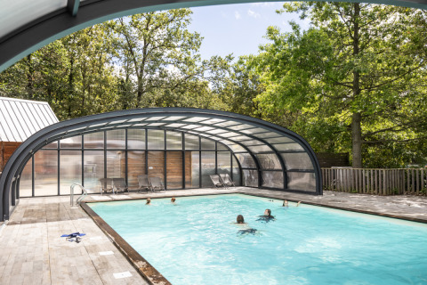 Piscina exterior con cubierta parcial y personas nadando, rodeada de árboles en Huttopia Versailles, Francia.