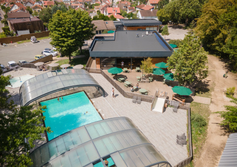 Vista aérea del parque vacacional Huttopia Versailles con piscina cubierta, terraza solárium y mesas exteriores.