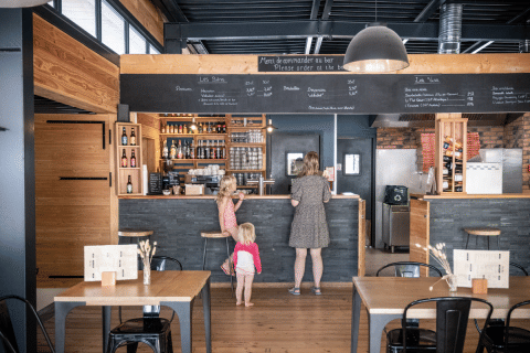 Een vrouw met twee kinderen plaatsen hun bestelling aan de bar van een restaurant in Huttopia Versailles, Île de France.