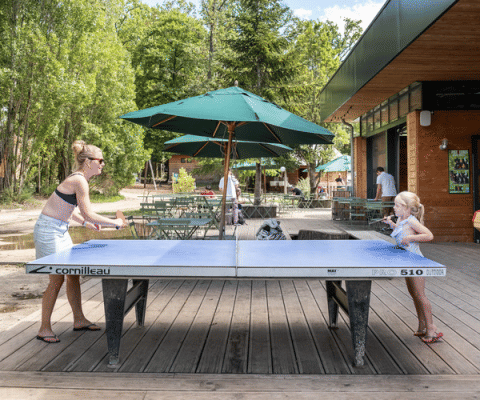 Two people play outdoor table tennis at Huttopia Versailles holiday park in Île de France, France, on a sunny day.