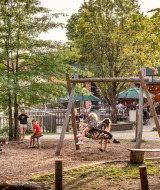 Children play on a playground swing as adults relax nearby at Huttopia Versailles holiday park in France.