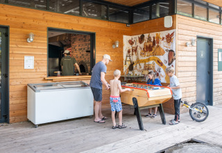 Kinder und Erwachsener spielen Tischfußball vor einer Holzhütte im Huttopia Versailles Ferienpark, Île-de-France.