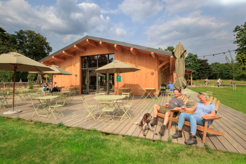 Mensen ontspannen op het terras bij Huttopia De Meinweg vakantiepark in Limburg, Nederland, met tafels en parasols.