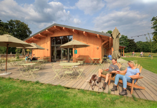 Menschen entspannen auf der Terrasse einer Holzhütte mit Hund in Huttopia De Meinweg, Limburg, Niederlande.
