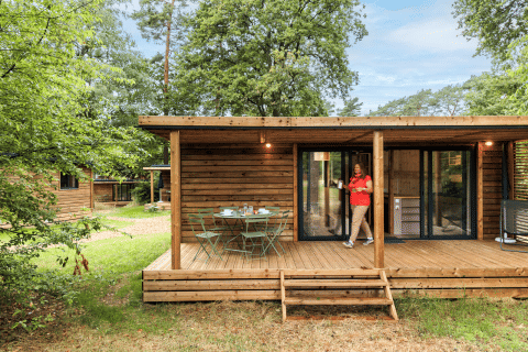 Cabaña de madera con terraza y mesa de jardín en Huttopia De Meinweg, Limburg, Países Bajos, rodeada de naturaleza.