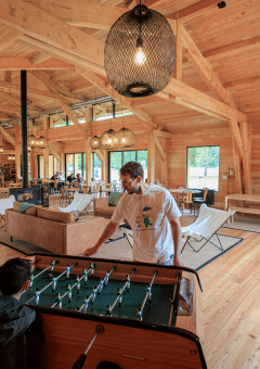 Interior de madera en área común de Huttopia De Meinweg, personas juegan futbolín y se relajan.