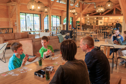 Familias jugando a las cartas y relajándose en una sala común acogedora en Huttopia De Meinweg, Limburg.