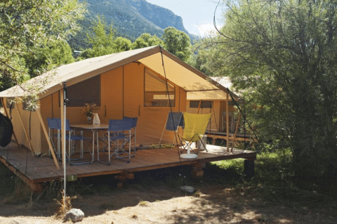 Ein Bungalowzelt Classic Safari-Zelt mit Holzdeck, Campingtisch und Stühlen in grüner Umgebung.