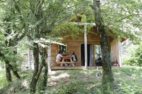 To personer sidder ved et bord foran Chalet Montana i Huttopia Vallouise, Frankrig, omgivet af grønne træer.