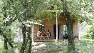 To personer sidder ved et bord foran Chalet Montana i Huttopia Vallouise, Frankrig, omgivet af grønne træer.