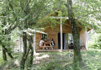 To personer sidder ved et bord foran Chalet Montana i Huttopia Vallouise, Frankrig, omgivet af grønne træer.
