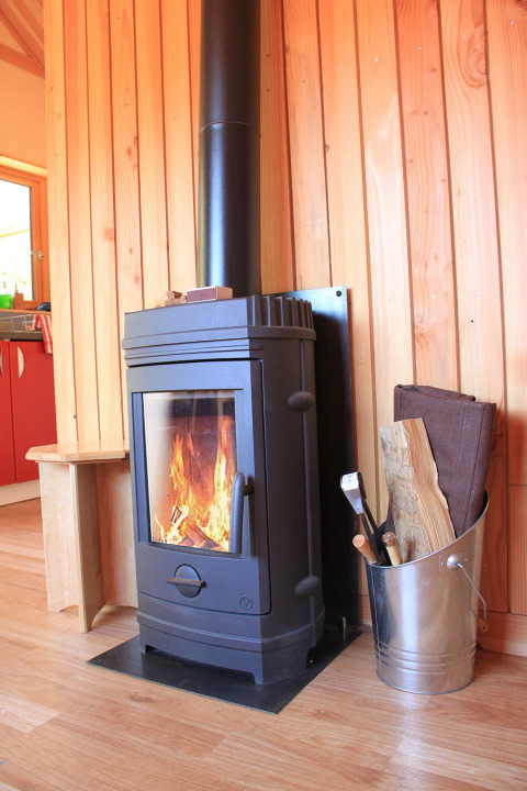 Estufa de leña encendida junto a cubo con leña y pared de madera en Chalet Montana, Huttopia Vallouise.