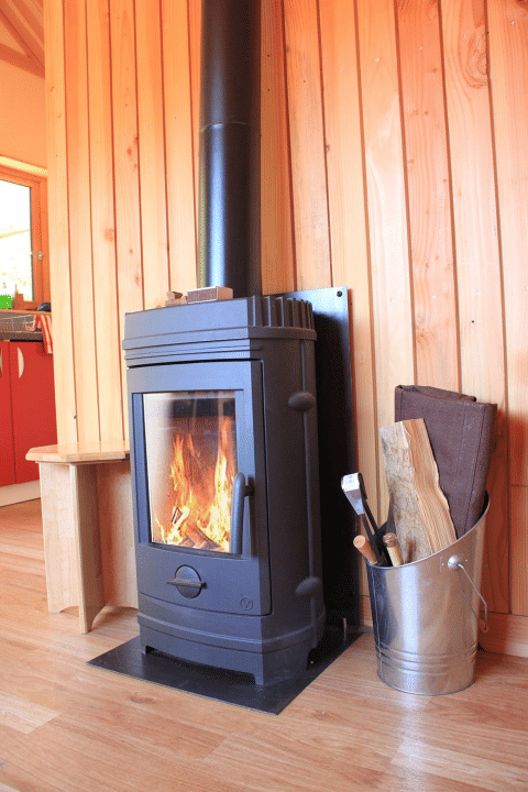 Estufa de leña encendida junto a cubo con leña y pared de madera en Chalet Montana, Huttopia Vallouise.
