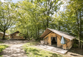 Safari tent Trappeur op een camping tussen groene bomen, met houten terras en buitenmeubilair.