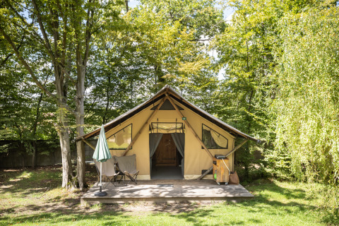 Tienda safari Trappeur en Huttopia Versailles, Francia, rodeada de árboles y mobiliario exterior.