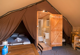 Interior de una tienda safari con cama doble, baño, mesa de comedor y estantería de cocina con platos.