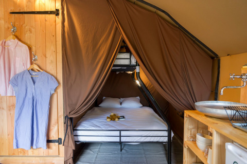 Interior de una tienda safari con cama doble, litera, ropa colgando y una pequeña cocina con fregadero.