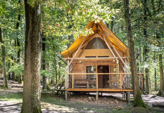 Cabin named Cahutte with yellow roof in a lush green forest, surrounded by tall trees and sunlight.