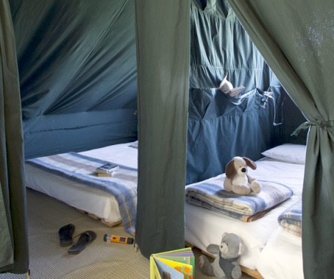 Inside a Cahutte at Huttopia Versailles, France, featuring two beds with blankets, toys, and a cozy camping setup.