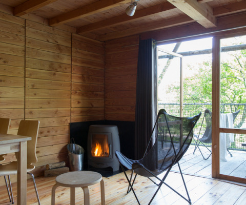Interior de cabaña de madera en Cahutte de Huttopia Versailles, Francia, con chimenea y terraza acristalada.