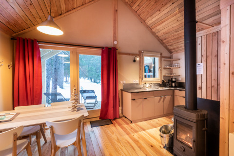 Sfeervolle glampinghut van hout met open haard, eettafel en uitzicht op een besneeuwd bos buiten.