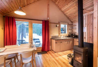 Acogedora cabaña glamping con decoración de madera, estufa, mesa de comedor y vistas al bosque nevado.