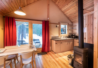 Intérieur de cabane glamping chaleureuse en bois avec poêle, table à manger et vue sur la forêt enneigée.