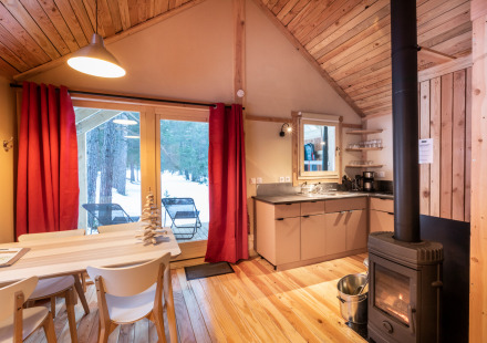 Sfeervolle glampinghut van hout met open haard, eettafel en uitzicht op een besneeuwd bos buiten.