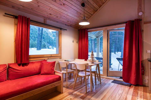 Knusse glampingaccommodatie met rode gordijnen, houten meubels en uitzicht op een besneeuwd bos door ramen.