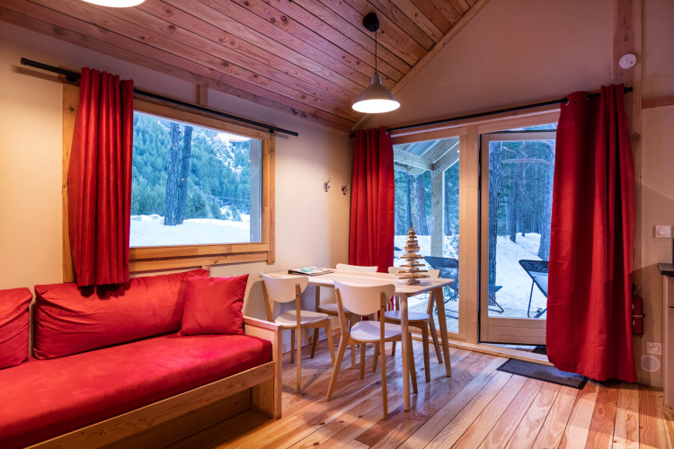 Acogedora cabaña glamping con cortinas rojas, sofá y vistas nevadas a un bosque a través de ventanas grandes.