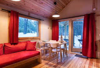 Acogedora cabaña glamping con cortinas rojas, sofá y vistas nevadas a un bosque a través de ventanas grandes.