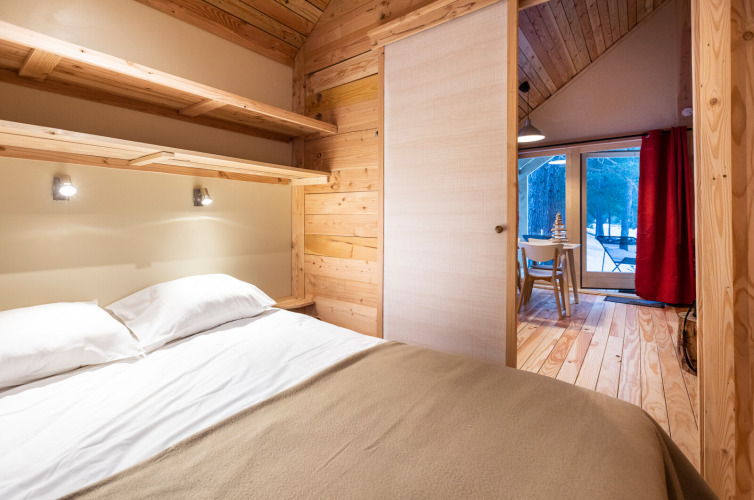 Acogedora habitación de glamping con cama doble, paredes de madera y vista al bosque por grandes ventanas.
