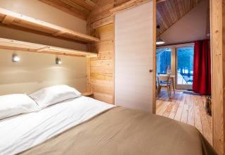 Acogedora habitación de glamping con cama doble, paredes de madera y vista al bosque por grandes ventanas.