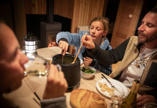 Familie genießt ein Fondue-Abendessen in einer gemütlichen Holzhütte mit Kamin in Huttopia la Clarée, Frankreich.