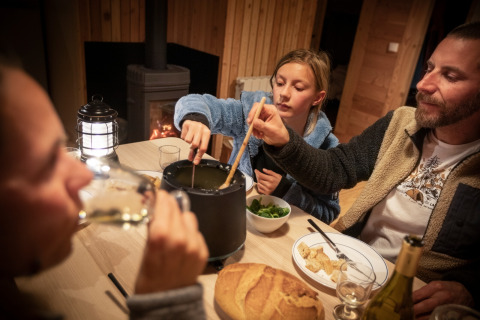 Famiglia che gusta una cena a base di fonduta davanti al camino in uno chalet di legno a Huttopia la Clarée, Francia.