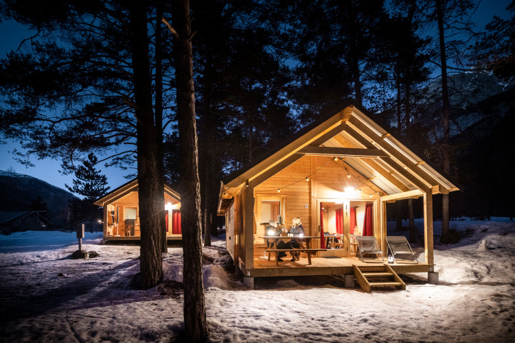 Cozy wooden cabins lit up in a snowy forest evening at Chalet Montana, Huttopia la Clarée, France.