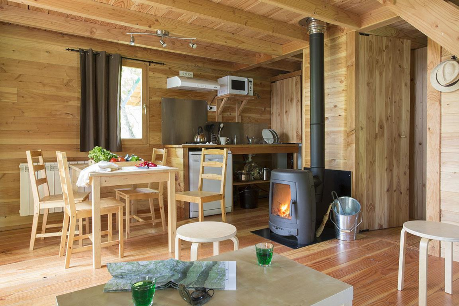 Gezellige houten cabin-interieur met eettafel, houtkachel en kitchenette bij Huttopia Versailles, Frankrijk.