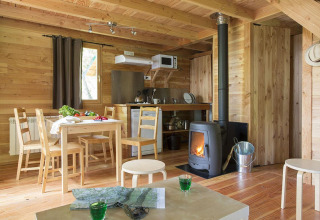 Intérieur d'une cabane en bois chaleureuse avec table, poêle et kitchenette à Huttopia Versailles, France.