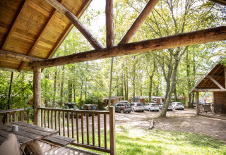 Blick von einer Holzhütte mit überdachter Terrasse bei Huttopia Versailles, umgeben von Autos und Bäumen.