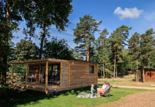 Houten Chalet Evasion tussen de bomen met twee volwassenen en een kind op het gras bij Huttopia De Meinweg.