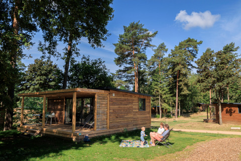 Chalet Evasion in legno immerso tra gli alberi, due adulti e un bambino si rilassano all'aperto a Huttopia De Meinweg.