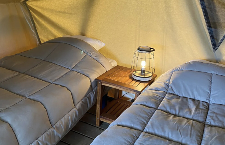 Zwei Einzelbetten mit grauen Decken und einer Laterne auf einem Holztisch in einem Safarizelt in Frankreich.