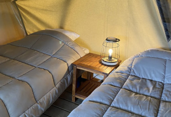 Dos camas individuales con edredones grises y una lámpara sobre una mesa de madera en una tienda safari en Francia.