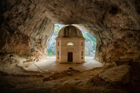 A small domed chapel stands inside a large cave near Sassoferrato, Marche, Italy, illuminated by sunlight.