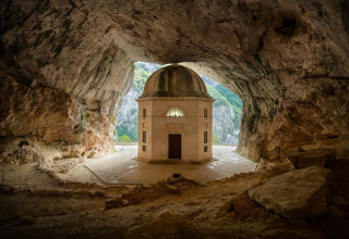A small domed chapel stands inside a large cave near Sassoferrato, Marche, Italy, illuminated by sunlight.