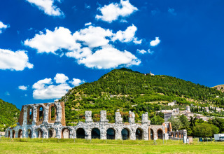 Ruins of an ancient Roman amphitheater surrounded by green hills near Sassoferrato, Marche, Italy under a blue sky.